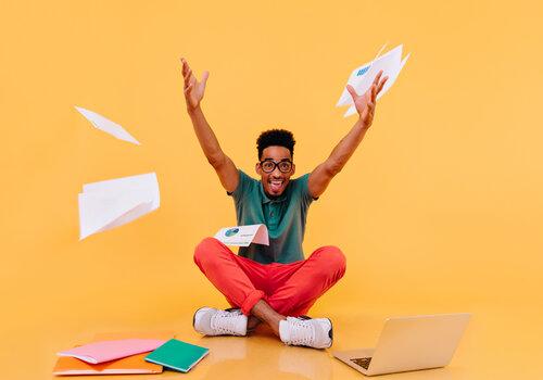 Les suprises de la science! Etudiant joyeux levant les bras, entouré de papiers, ordinateur...