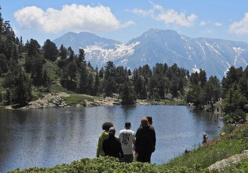 Cette photo montre un groupe de quatre jeunes, deux hommes et deux femmes, debout au bord d'un lac alpin entouré de montagnes enneigées et de forêts verdoyantes. Ils regardent le paysage, où se détachent les sommets des montagnes sous un ciel bleu parsemé de quelques nuages blancs.