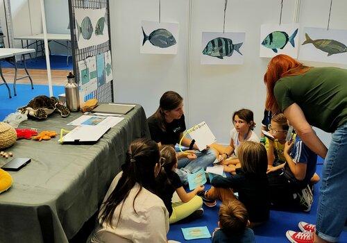Des enfants assis sur une moquette bleue regardent et écoutent une femme qui leur présente des poissons