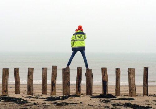À l'eau, la Terre ? Personne en veste jaune fluo marquée "ANPU", debout sur des pieux en bois sur une plage, face à la mer brumeuse.