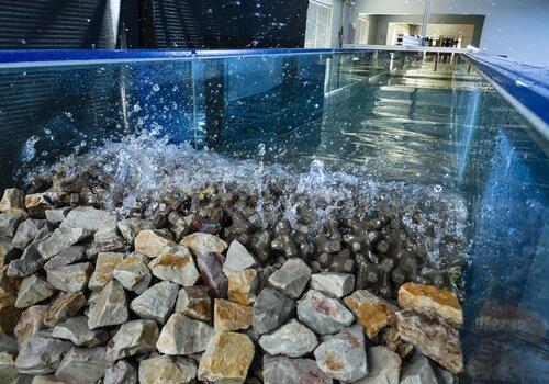 Texte alternatif concis :  > Expérience de houle dans un canal en verre rempli d'eau avec des blocs rocheux au fond.