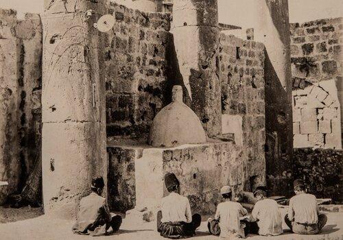 Photographie en noir et blanc prise à Palmyre, montrant un groupe d’enfants assis devant un four installé entre deux colonnes.
