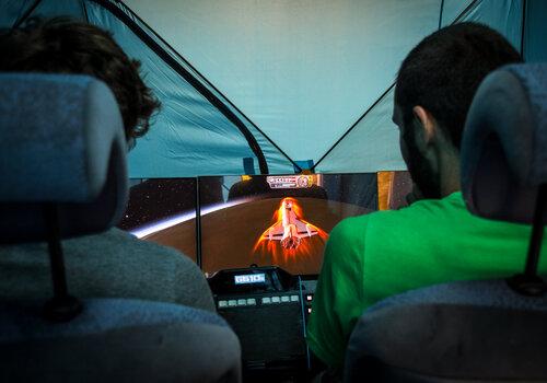 Embarquez dans la capsule ! Deux hommes sont assis, de dos, aux commandes d'un écran qui représente une fusée en vol.