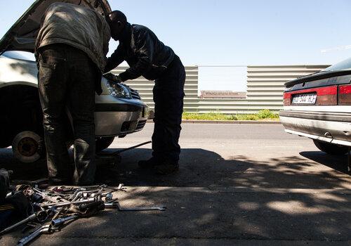 Deux garagistes dans la rue, en train de réparer une voiture capot ouvert. 