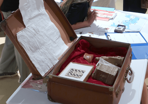 Une valise en cuir marron remplie d'objets archéologiques (statuette, morceau de céramique) posés sur du velours rouge. La valise est sur une table couverte de papiers.