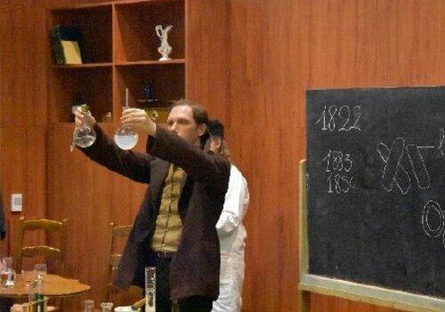 Un comédien interprète Louis Pasteur sur scène avec des ballons de chimie en verre dans les mains.