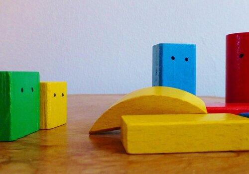 Photographie de plusieurs blocs de construction en bois colorés (vert, jaune, bleu, rouge) disposés sur une table en bois. Certains blocs sont décorés de deux petits points noirs évoquant des yeux, leur donnant l'apparence de petits personnages ou de molécules animées. Les formes sont variées : cubes, cylindres et arcs de cercle.