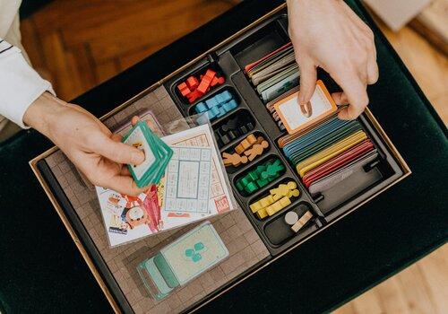 Deux mains tiennent des cartes au dessus d'une boîte de jeu remplie de pions et de cartes colorées