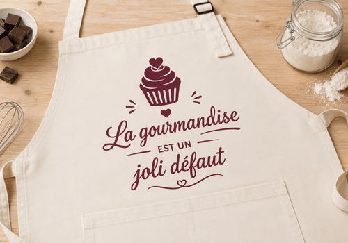 Tablier blanc sur une table avec l'inscription "La gourmandise est un joli défaut" et un dessin de cupcake