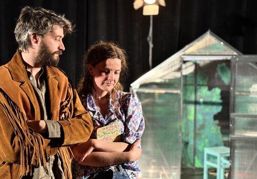 Visuel du spectacle : Un homme et une femme les bras croisés se trouvent devant une serre au second plan. 