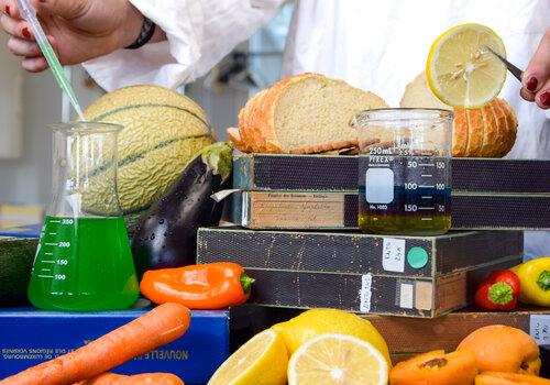 Une personne en blouse manipule des instruments de laboratoire au milieu de fruits, légumes, pain et autres aliments.