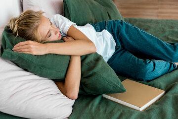 Une femme dort sur un lit, enlaçant un oreiller vert. Un livre fermé est posé à côté d'elle
