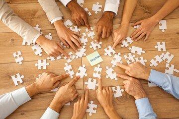 Des mains de couleurs différentes pointent vers un post-it sur lequel est écrit "solution". Des morceaux de puzzles blanc sont étalés au sol. 