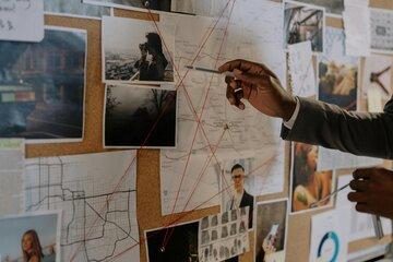 Sur un tableau en liège, sont épinglés une carte routière, un plan urbain, des empreintes digitales, un paysage sombre et brumeux avec une maison, des visages, et d’autres documents cartographiques. Plusieurs fils rouges sont tendus entre différents éléments. Une personne en costume (probablement un enquêteur) pointe un endroit avec un stylo, en essayant d’élaborer ou expliquer un lien. 