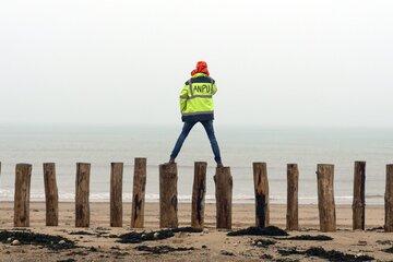 Personne en veste jaune fluo marquée "ANPU", debout sur des pieux en bois sur une plage, face à la mer brumeuse.