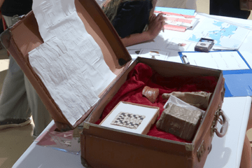 Une valise en cuir marron remplie d'objets archéologiques (statuette, morceau de céramique) posés sur du velours rouge. La valise est sur une table couverte de papiers.