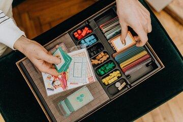 Deux mains tiennent des cartes au dessus d'une boîte de jeu remplie de pions et de cartes colorées