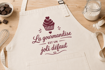 Tablier blanc sur une table avec l'inscription "La gourmandise est un joli défaut" et un dessin de cupcake