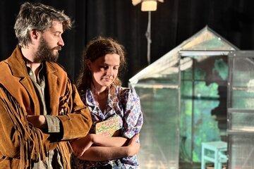 Visuel du spectacle : Un homme et une femme les bras croisés se trouvent devant une serre au second plan. 