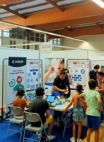 Les stands du Village des sciences Sur un stand du village des sciences de montpellier, deux femmes présentent à des enfants et une adulte des documents sur l'étude des sons et de l'audition humaine