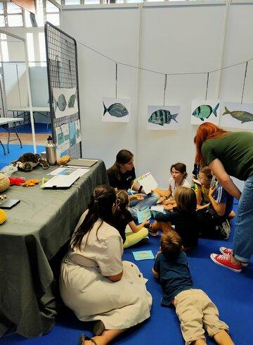Des enfants assis sur une moquette bleue regardent et écoutent une femme qui leur présente des poissons